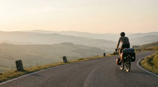 Cycliste avec vélo chargé de sacoches traversant un paysage européen vallonné au lever du soleil